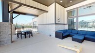 This well-appointed covered outdoor patio offers a versatile and inviting space designed for both relaxation and entertainment. The area is defined by a clean, contemporary architectural style, featuring board-and-batten siding in crisp white and a lower accent section clad in light-toned stone, lending the space a polished and modern aesthetic.

The patio is partially covered by a structured overhead canopy with recessed lighting, providing shade and ambiance while remaining open to the outdoors. The flooring consists of a smooth, neutral-toned concrete surface that extends throughout the entire area.

The space is organized into distinct functional zones. A comfortable lounge area is anchored by a dark wicker sectional sofa dressed in bold blue cushions, ideal for casual gatherings. Adjacent to this is an outdoor dining setup featuring a round table with black chairs, well-suited for al fresco meals. A cornhole board is also present, adding a recreational element to the space.

A black wrought iron fence borders the perimeter, offering a sense of enclosure while preserving open views of the surrounding neighborhood. Natural light flows generously into the space, enhancing its bright and airy atmosphere. Overall, this patio presents a functional and stylish outdoor living environment suitable for a range of social and leisure activities.