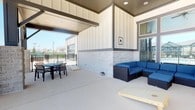 This well-appointed covered outdoor patio offers a versatile and inviting space designed for both relaxation and entertainment. The area is defined by a clean, contemporary architectural style, featuring board-and-batten siding in crisp white and a lower accent section clad in light-toned stone, lending the space a polished and modern aesthetic.

The patio is partially covered by a structured overhead canopy with recessed lighting, providing shade and ambiance while remaining open to the outdoors. The flooring consists of a smooth, neutral-toned concrete surface that extends throughout the entire area.

The space is organized into distinct functional zones. A comfortable lounge area is anchored by a dark wicker sectional sofa dressed in bold blue cushions, ideal for casual gatherings. Adjacent to this is an outdoor dining setup featuring a round table with black chairs, well-suited for al fresco meals. A cornhole board is also present, adding a recreational element to the space.

A black wrought iron fence borders the perimeter, offering a sense of enclosure while preserving open views of the surrounding neighborhood. Natural light flows generously into the space, enhancing its bright and airy atmosphere. Overall, this patio presents a functional and stylish outdoor living environment suitable for a range of social and leisure activities.