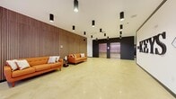 The Keys is a thoughtfully designed building lobby that makes a strong first impression through its contemporary aesthetic and generous proportions. The space features polished concrete flooring that reflects light throughout, contributing to a bright and open atmosphere. A dramatic full-height slatted wood accent wall anchors the lounge area, adding warmth and texture against the otherwise clean, modern palette of white and dark charcoal finishes. A seating arrangement composed of bold orange mid-century-style sofas and a small side table provides a comfortable waiting area for residents and guests. On the opposite side of the lobby, two stainless steel elevator doors are set within a dark-framed alcove, offering easy vertical access throughout the building. Cylindrical black pendant lights are distributed evenly across the ceiling, providing focused illumination that complements the polished floor below. A prominently branded feature wall displaying "The Keys" signage serves as a striking focal point near the entrance. Floor-to-ceiling black-framed windows line one side of the lobby, inviting abundant natural light and offering views of the surrounding streetscape. An exposed concrete structural column adds an industrial character that balances the refined finishes throughout the space.