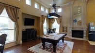 This sophisticated entertainment room is defined by its dramatic double-height ceilings, which are lined with a row of clerestory windows that bathe the space in natural light alongside larger standard windows dressed in flowing gold drapes. The warm, textured walls in a rich ochre tone lend a stately ambiance throughout. At the center of the room sits a full-size billiard table in dark wood, anchored by an ornate pendant light fixture that doubles as a ceiling fan — a functional yet striking focal point. Rich hardwood floors extend across the entire space, partially covered by a detailed red and cream area rug that complements the room's warm color palette. A wall-mounted flat-screen television is positioned above a dark wood media console along one wall, providing a secondary entertainment option. A dedicated cue rack nearby offers convenient storage for billiard accessories. An elegant white marble fireplace with decorative columns adds a classical architectural touch to one corner. A small dining or card table with chairs is positioned near the window, making excellent use of the natural light. The overall ambiance is refined yet inviting, combining classical design elements with modern entertainment amenities.