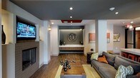 This single-floor space presents a sleek and contemporary design aesthetic that immediately conveys a modern, urban sensibility. The open layout centers around a well-appointed reception or leasing desk area, set against a deep-toned feature wall adorned with a large decorative "@" symbol — a bold nod to connected, modern living. Recessed lighting and a tiered ceiling with warm orange accents add architectural interest overhead, while rich hardwood-style flooring grounds the space with warmth and texture. A lounge area to one side features a comfortable gray sofa accented with vibrant orange throw pillows, paired with a rustic wooden coffee table and industrial-style sculptural décor pieces that add character and depth. A wall-mounted flat-screen television and a classic fireplace surround on the adjacent wall enhance the welcoming, residential feel of the communal area. Warm sconce lighting complements the recessed fixtures, creating a balanced and inviting ambiance throughout. Colorful artwork on the walls adds visual energy, tying together the gray, white, and orange color palette that defines the space. Overall, this thoughtfully designed interior blends functional purpose with sophisticated style.