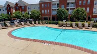 This outdoor amenity space centers around a generously sized swimming pool with crystal-clear blue water, ranging from a shallow end to a deeper section, clearly marked for safety. The pool's clean geometric shape is complemented by a wide surrounding deck finished in a combination of brick-toned coping and light concrete, providing ample space for lounging and movement.

The sun deck is well-furnished with multiple cushioned lounge chairs and side tables, arranged to take full advantage of the open-air setting and natural sunlight. Lush mature trees line the perimeter, offering pockets of shade and a sense of natural enclosure within the otherwise open space.

A sturdy wrought iron fence borders the pool area, providing both security and a clear boundary while maintaining visibility to the surrounding landscape. The backdrop features classic multi-story red brick buildings with arched windows, balconies, and traditional architectural detailing, contributing to a cohesive and upscale residential aesthetic.

The overall ambiance is inviting and resort-like, designed to encourage relaxation and outdoor enjoyment. The combination of well-maintained landscaping, quality furnishings, and thoughtful layout makes this outdoor pool area a standout communal amenity.