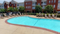 This outdoor amenity space centers around a generously sized swimming pool with crystal-clear blue water, ranging from a shallow end to a deeper section, clearly marked for safety. The pool's clean geometric shape is complemented by a wide surrounding deck finished in a combination of brick-toned coping and light concrete, providing ample space for lounging and movement.

The sun deck is well-furnished with multiple cushioned lounge chairs and side tables, arranged to take full advantage of the open-air setting and natural sunlight. Lush mature trees line the perimeter, offering pockets of shade and a sense of natural enclosure within the otherwise open space.

A sturdy wrought iron fence borders the pool area, providing both security and a clear boundary while maintaining visibility to the surrounding landscape. The backdrop features classic multi-story red brick buildings with arched windows, balconies, and traditional architectural detailing, contributing to a cohesive and upscale residential aesthetic.

The overall ambiance is inviting and resort-like, designed to encourage relaxation and outdoor enjoyment. The combination of well-maintained landscaping, quality furnishings, and thoughtful layout makes this outdoor pool area a standout communal amenity.