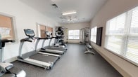 Located at 300 Bellamy Ave in Conway, South Carolina, this single-story amenity center is designed to support an active and connected lifestyle. The facility's centerpiece is a spacious, well-lit fitness room featuring rubber-floored workout areas bathed in natural light from multiple large windows fitted with white blinds. The gym is outfitted with a comprehensive selection of TRUE Fitness cardio equipment, including treadmills, upright bikes, and an elliptical, alongside a multi-station cable machine, a leg press unit, and a TRX suspension training system. A wall-mounted television and ceiling fan keep the environment comfortable during workouts. Free weights and a full dumbbell rack, plus medicine balls and stability balls on a dedicated storage rack, round out the strength training options. A separate, private room houses a standalone tanning booth, providing a convenient self-care amenity. Adjacent to the fitness area is a bright business lounge featuring a granite-topped counter with multiple iMac workstations, framed whiteboards, and bar-height seating — ideal for remote work or study. The room benefits from tall ceilings and generous window light. Throughout, the facility maintains a clean, modern aesthetic with neutral warm-white walls and light wood-tone flooring in the lounge areas, presenting a polished and functional environment.