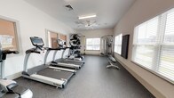 Located at 300 Bellamy Ave in Conway, South Carolina, this single-story amenity center is designed to support an active and connected lifestyle. The facility's centerpiece is a spacious, well-lit fitness room featuring rubber-floored workout areas bathed in natural light from multiple large windows fitted with white blinds. The gym is outfitted with a comprehensive selection of TRUE Fitness cardio equipment, including treadmills, upright bikes, and an elliptical, alongside a multi-station cable machine, a leg press unit, and a TRX suspension training system. A wall-mounted television and ceiling fan keep the environment comfortable during workouts. Free weights and a full dumbbell rack, plus medicine balls and stability balls on a dedicated storage rack, round out the strength training options. A separate, private room houses a standalone tanning booth, providing a convenient self-care amenity. Adjacent to the fitness area is a bright business lounge featuring a granite-topped counter with multiple iMac workstations, framed whiteboards, and bar-height seating — ideal for remote work or study. The room benefits from tall ceilings and generous window light. Throughout, the facility maintains a clean, modern aesthetic with neutral warm-white walls and light wood-tone flooring in the lounge areas, presenting a polished and functional environment.