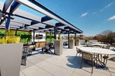 Located at 1350 Dresden Drive NE in Brookhaven, Georgia, this rooftop terrace offers a well-appointed outdoor amenity space designed for both relaxation and social gatherings. The large-format porcelain tile deck extends across a generous open-air footprint, providing ample room for a variety of seating configurations, including round dining tables with chairs, chaise lounges for sunbathing, and additional lounge furniture arranged throughout the space.

A standout feature is the modern pergola structure situated along one side of the terrace. Constructed with sleek steel framing and retractable or louvered roof panels, it provides shade and shelter while maintaining an open, airy feel. Beneath the pergola, a dedicated entertainment zone includes an outdoor television, a bar-height counter with bar seating, lush vertical greenery, and large planter boxes adding organic texture to the contemporary design.

The open portion of the terrace is bathed in natural sunlight and offers sweeping views of the Brookhaven neighborhood skyline and surrounding tree canopy. Low perimeter walls and railing systems ensure safety while preserving sightlines. Manicured topiary accents and tall rectangular planters are distributed throughout, contributing a refined, curated aesthetic. The overall design balances modern architectural elements with functional outdoor living, making it a versatile amenity space suited for both quiet relaxation and social events.