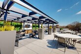 Located at 1350 Dresden Drive NE in Brookhaven, Georgia, this rooftop terrace offers a well-appointed outdoor amenity space designed for both relaxation and social gatherings. The large-format porcelain tile deck extends across a generous open-air footprint, providing ample room for a variety of seating configurations, including round dining tables with chairs, chaise lounges for sunbathing, and additional lounge furniture arranged throughout the space.

A standout feature is the modern pergola structure situated along one side of the terrace. Constructed with sleek steel framing and retractable or louvered roof panels, it provides shade and shelter while maintaining an open, airy feel. Beneath the pergola, a dedicated entertainment zone includes an outdoor television, a bar-height counter with bar seating, lush vertical greenery, and large planter boxes adding organic texture to the contemporary design.

The open portion of the terrace is bathed in natural sunlight and offers sweeping views of the Brookhaven neighborhood skyline and surrounding tree canopy. Low perimeter walls and railing systems ensure safety while preserving sightlines. Manicured topiary accents and tall rectangular planters are distributed throughout, contributing a refined, curated aesthetic. The overall design balances modern architectural elements with functional outdoor living, making it a versatile amenity space suited for both quiet relaxation and social events.
