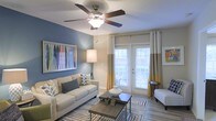 This single-story space features a well-appointed living area that blends comfort and style. The room is anchored by a striking blue accent wall, which provides a bold contrast to the neutral beige tones found throughout. A ceiling fan with an integrated light fixture is centrally positioned, offering both functionality and a finishing architectural touch.

Natural light flows generously into the space through a set of French doors dressed with floor-length gold and white curtains, which also provide access to an outdoor area. The wood-look flooring adds warmth underfoot, while a patterned area rug defines the seating zone and introduces additional visual interest.

The living area is thoughtfully arranged, with ample room for seating and a central coffee table. Framed artwork decorates the walls, contributing to the curated, cohesive aesthetic of the space. A floor lamp provides supplemental ambient lighting, enhancing the room's inviting atmosphere.

Overall, the space presents a clean, modern aesthetic that balances practical living with stylish design elements, making it a comfortable and visually appealing environment.