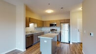 This single-floor residence offers a thoughtfully designed open-concept layout that seamlessly connects the kitchen and living areas. The kitchen serves as the heart of the space, showcasing warm medium-tone wood cabinetry that extends to the upper walls for ample storage. Granite-style countertops provide generous prep space and are complemented by a freestanding center island, ideal for casual dining or additional workspace. A full suite of stainless steel appliances — including a refrigerator, range with microwave above, and dishwasher — adds a modern, functional touch. The sink is positioned along the perimeter countertop beneath upper cabinetry, keeping the layout efficient and practical. Durable hardwood-style flooring runs throughout the main living and kitchen areas, lending a cohesive, clean aesthetic. The living area is open and well-lit, with neutral wall tones that create a calm, versatile backdrop. The bedroom features plush carpeting and a window fitted with blinds that allows for natural light and outdoor views. The bathroom is finished with wood-look tile flooring and includes a combined tub and shower unit with chrome fixtures, maintaining a clean and functional design throughout.