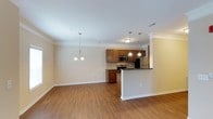 This single-story residence at 150 Horizon Trl in Garner, North Carolina presents an open-concept floor plan that connects the living, dining, and kitchen areas into one cohesive, light-filled space. Crown molding runs along the ceiling throughout, adding a refined architectural detail that complements the neutral, warm-toned walls and crisp white trim.

The kitchen is appointed with rich medium-toned wood cabinetry, dark countertops, and stainless steel appliances, including a microwave, range, and refrigerator. A raised peninsula serves as a natural divider between the kitchen and the adjoining living and dining areas, offering both additional counter space and a casual seating option. Pendant lighting above the peninsula provides focused task illumination, while a chandelier in the dining area adds an elegant finishing touch.

The living and dining spaces feature continuous wood-look hard-surface flooring that extends throughout, providing a warm and cohesive aesthetic that is easy to maintain. The open layout allows for flexible furniture arrangement and promotes a sense of spaciousness. Overall, the space maintains a clean, welcoming ambiance well-suited for everyday living.