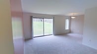 This single-floor residence offers a clean, understated aesthetic defined by neutral wall tones and soft, plush wall-to-wall carpeting throughout. The living area serves as the heart of the space, where a large sliding glass door commands attention as the focal point of the room. This expansive glass feature floods the interior with abundant natural light and provides direct views of a lush, green landscaped yard, creating a seamless connection between the indoors and outdoors. A secondary vertical window alongside the sliding door further enhances the brightness of the room. The space is complemented by a modest pendant light fixture that adds a subtle decorative touch to the ceiling. The overall layout feels open and airy, with clean wall lines and a simple, functional design that provides a versatile canvas for a variety of interior styles. The neutral color palette of warm beige and off-white tones throughout contributes to a calm and welcoming ambiance. The single-floor layout ensures convenient, accessible living, making the space practical and easy to navigate. Overall, this residence balances simplicity and comfort, with its standout natural light and outdoor connectivity being its most defining characteristics.