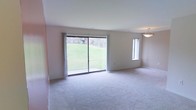 This single-floor residence offers a clean, understated aesthetic defined by neutral wall tones and soft, plush wall-to-wall carpeting throughout. The living area serves as the heart of the space, where a large sliding glass door commands attention as the focal point of the room. This expansive glass feature floods the interior with abundant natural light and provides direct views of a lush, green landscaped yard, creating a seamless connection between the indoors and outdoors. A secondary vertical window alongside the sliding door further enhances the brightness of the room. The space is complemented by a modest pendant light fixture that adds a subtle decorative touch to the ceiling. The overall layout feels open and airy, with clean wall lines and a simple, functional design that provides a versatile canvas for a variety of interior styles. The neutral color palette of warm beige and off-white tones throughout contributes to a calm and welcoming ambiance. The single-floor layout ensures convenient, accessible living, making the space practical and easy to navigate. Overall, this residence balances simplicity and comfort, with its standout natural light and outdoor connectivity being its most defining characteristics.