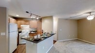This single-floor space showcases an open-concept layout that seamlessly connects the kitchen and living area. The kitchen is well-appointed with warm honey-toned wood cabinetry providing ample storage, complemented by dark granite countertops that add a touch of contrast and sophistication. A peninsula-style breakfast bar extends into the space, creating a natural boundary between the kitchen and living area while offering additional seating and prep space. White appliances, including a range and refrigerator, are neatly integrated beneath the cabinetry, and an under-cabinet range hood adds a functional touch. Track lighting mounted on the ceiling illuminates the kitchen efficiently, highlighting the workspace below. The transition into the living area is marked by a shift from hardwood-style flooring in the kitchen to plush carpeting, contributing to a warm and comfortable ambiance. Neutral beige tones on the walls throughout create a cohesive and inviting atmosphere. A doorway visible in the background suggests additional rooms beyond, adding to the functional layout of the space.