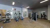 This fitness center offers a thoughtfully designed workout space spread across a single floor. The room features neutral gray walls paired with textured, patterned carpeting that adds warmth underfoot. Overhead, flush-mounted fluorescent lighting ensures the space is well-lit throughout.

The cardio section is anchored by multiple elliptical machines, each equipped with individual screens for entertainment during workouts. Wall-mounted televisions are positioned at an elevated level, providing additional viewing options across the room. Large windows along one wall invite natural light and contribute to an open, airy atmosphere.

On the strength training side, a multi-functional cable machine serves as the centerpiece, supplemented by additional equipment visible toward the back of the room. Full-length mirrors line the walls in the strength area, enhancing the sense of space while allowing users to monitor their form.

Glass-paneled double doors provide access to an adjacent area, suggesting additional fitness or amenity spaces nearby. Overall, the fitness center maintains a clean, modern aesthetic with a practical layout designed to accommodate a variety of workout routines comfortably.