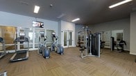 This fitness center offers a thoughtfully designed workout space spread across a single floor. The room features neutral gray walls paired with textured, patterned carpeting that adds warmth underfoot. Overhead, flush-mounted fluorescent lighting ensures the space is well-lit throughout.

The cardio section is anchored by multiple elliptical machines, each equipped with individual screens for entertainment during workouts. Wall-mounted televisions are positioned at an elevated level, providing additional viewing options across the room. Large windows along one wall invite natural light and contribute to an open, airy atmosphere.

On the strength training side, a multi-functional cable machine serves as the centerpiece, supplemented by additional equipment visible toward the back of the room. Full-length mirrors line the walls in the strength area, enhancing the sense of space while allowing users to monitor their form.

Glass-paneled double doors provide access to an adjacent area, suggesting additional fitness or amenity spaces nearby. Overall, the fitness center maintains a clean, modern aesthetic with a practical layout designed to accommodate a variety of workout routines comfortably.