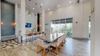 This well-appointed community clubhouse is designed to foster social interaction and relaxation across a single, thoughtfully organized floor. The space immediately impresses with its soaring ceilings, wide-plank wood-look tile flooring, and floor-to-ceiling windows that flood the interior with natural light and offer views of lush tropical landscaping and palm trees outside.

The main gathering area serves as the heart of the clubhouse, featuring a large communal dining table surrounded by woven rattan-style chairs, complemented by a striking herringbone-patterned teal tile accent wall, pendant lighting, and a built-in bar-height counter with leather barstools. A wall-mounted television and decorative sconce lighting complete the space.

Adjacent to the dining area, an expansive lounge and game room offers plush seating, globe pendant light fixtures, and multiple billiard tables, all framed by large windows overlooking the outdoor greenery. A separate, more intimate lounge nook provides a quiet retreat with swivel armchairs and a wall-mounted television set against a textured vertical panel accent wall.

Throughout, the design is unified by a coastal-contemporary aesthetic, featuring abstract blue-toned artwork, aqua accent pillows, and a bright, airy color palette that creates an inviting and sophisticated communal environment.