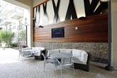 This covered outdoor amenity space is a thoughtfully designed communal lounge nestled within the courtyard of a multi-story residential complex. The space features a striking architectural ceiling with exposed beams and bold geometric wall art rendered in black and white, adding a contemporary artistic flair to the environment.

The flooring throughout is laid in classic brick pavers, lending a warm, textured character to the space. One wall showcases a rich horizontal wood panel backdrop, beneath which a flat-screen television is mounted — ideal for gatherings and communal viewing. Below the screen, a stacked stone base supports a long marble-topped bar counter with accompanying seating, complemented by a stainless steel mini-fridge unit for added convenience.

The lounge area is furnished with comfortable wicker sectional sofas and accent tables, creating an inviting setting for relaxation and socializing. Elegant Tiffany-style floor lamps add a warm decorative glow throughout the space.

Opening directly onto the broader courtyard, the covered pavilion enjoys an abundance of natural light and seamless views of the surrounding landscaped grounds, outdoor dining areas, and the multi-story building facades with their balconied units. The overall ambiance is modern yet welcoming, blending upscale design details with practical amenities suited for community living.