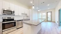 This single-floor residence showcases a thoughtfully designed open-concept layout that seamlessly connects the kitchen and living areas. The kitchen serves as the focal point, featuring a large granite-topped island with built-in storage and pendant lighting overhead. White upper cabinetry contrasts with warm wood-toned lower cabinets, while a white subway tile backsplash adds a clean, contemporary detail. A full suite of stainless steel appliances includes a side-by-side refrigerator with ice and water dispenser, electric range, built-in microwave, and dishwasher. Light hardwood-style flooring extends throughout the main living areas, contributing to a bright and cohesive aesthetic. Large windows fitted with horizontal blinds allow natural light to filter through the living space, which also features access to an outdoor area via a glass door. The bedroom areas offer plush carpeting in at least one room and hardwood-style flooring in another, each with windows providing natural light and outdoor views. The overall design palette is neutral and contemporary, emphasizing clean lines, modern finishes, and a bright, airy atmosphere throughout.