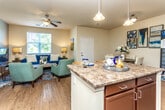 This single-floor residence offers a thoughtfully designed open-concept layout that seamlessly connects the kitchen, dining, and living areas. The kitchen is equipped with rich dark-wood cabinetry, granite-style countertops, black appliances including a range, built-in microwave, dishwasher, and refrigerator, along with a peninsula that provides additional prep space and casual seating. Track lighting and pendant fixtures illuminate the area effectively. The adjacent dining space accommodates a round table and chairs beneath a gallery wall arrangement, while the living room features hardwood-style flooring, a ceiling fan, and a window offering outdoor views. A second seating area functions as a media room with neutral-toned walls and direct access to the bedrooms. The bedrooms are carpeted and well-proportioned, each with windows that bring in natural light. One bedroom features dual nightstands and accent wall coloring, adding visual interest. The bathroom includes a granite-topped vanity with dark cabinetry, a tub-shower combination, and tile surround. Neutral wall tones and consistent finishes throughout contribute to a cohesive and comfortable living environment.