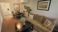 This two-floor residence offers a comfortable and well-appointed living experience, thoughtfully designed with a cohesive, earthy color palette throughout. The main living area is anchored by a plush sofa and a sleek dark wood coffee table, creating an inviting space for relaxation. Framed artwork adorns the neutral-toned walls, adding warmth and character to the room. The living space flows seamlessly into a dedicated dining area, where a round dark wood table is paired with matching cross-back chairs, ideal for everyday meals or entertaining. A tall indoor plant adds a natural, refreshing element to the shared space, enhancing the overall ambiance. The flooring features rich hardwood-style surfaces that complement the dark furnishings and warm wall tones, tying the design together cohesively. The layout makes efficient use of space across both floors, offering a practical and stylish environment. Crown molding details and recessed lighting contribute subtle architectural refinement to the interior. Overall, the space presents a classic, timeless aesthetic with a focus on comfort, functionality, and warm, welcoming design elements throughout.