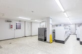 Located at 40 Tuxedo Crt in Toronto, this shared laundry facility offers a well-organized, functional space designed to serve building residents efficiently. The room features a generous open floor plan finished with durable terrazzo flooring throughout, providing a clean and easy-to-maintain surface. Bright fluorescent strip lighting runs along the textured white ceiling, ensuring the entire space is well-lit at all times.

The laundry area is equipped with multiple front-loading and top-loading washing machines and dryers arranged in rows, offering ample capacity for simultaneous use. A long countertop runs along one wall, featuring a double stainless steel utility sink with dark granite-style counters and white cabinetry below, providing a convenient area for hand-washing or sorting laundry. White subway tile accents the wall behind the counter, adding a clean, finished look.

A bulletin board near the entrance displays important notices and building information for residents. An open-area section of the room provides additional space, currently set up with a folding table and a payment kiosk for machine use. Exposed ductwork and baseboard heating are visible, and small ground-level windows allow a touch of natural light into the space. The facility also includes a fire safety station and clearly posted operating instructions throughout.