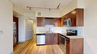 Located at 1000 Minor Ave in Seattle's First Hill neighborhood, this efficiently designed single-floor unit offers a well-organized layout that maximizes every square foot. The open-concept kitchen is a standout feature, outfitted with rich warm-toned wood cabinetry, light stone countertops, a classic white subway tile backsplash, and a full suite of stainless steel appliances including a range, microwave, and dishwasher. Track lighting overhead adds a modern, functional touch. Light wood flooring runs seamlessly throughout the entire unit, lending a cohesive and clean aesthetic. The living area is bright and airy, benefiting from a large picture window that frames views of the surrounding Seattle skyline, complemented by a ceiling fan for added comfort. The bedroom is a quiet, neutral space featuring a sliding-door closet with built-in shelving, offering practical storage. A hallway connects the main living spaces to the full bathroom, which is finished with a soaking tub and shower combo, a wood-accented vanity with open shelving, and matching light flooring. The overall design is clean and contemporary, with a neutral palette that creates a calm, inviting atmosphere throughout.