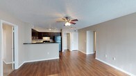 This single-floor residence offers a well-designed open-concept layout that seamlessly connects the living area and kitchen, creating a spacious and functional living environment. The living area features warm hardwood-style flooring that extends throughout, complemented by neutral beige walls and crisp white baseboards that give the space a clean, inviting aesthetic. A ceiling fan with an integrated light fixture and additional track lighting provide ample illumination across the open floor plan.

The kitchen is defined by rich, dark espresso-toned cabinetry paired with dark countertops, creating a bold contrast against the lighter surrounding walls. Black appliances, including a refrigerator and range, reinforce the kitchen's cohesive, modern look. A breakfast bar peninsula separates the kitchen from the living space, offering both a casual dining option and an additional workspace.

A textured popcorn ceiling adds dimension overhead, while recessed doors visible from the main living area lead to additional rooms, suggesting a practical and well-organized floor plan. The open sightlines throughout the main living space contribute to an airy, connected feel, making the residence feel welcoming and easy to navigate. Overall, the space balances contemporary finishes with functional design.