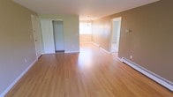 This single-floor space presents a clean, functional layout highlighted by light-toned hardwood flooring that runs throughout, lending warmth and continuity to the interior. The walls are finished in neutral earth tones — a soft sage green on one side transitioning to a warm taupe — creating a calm and welcoming atmosphere.

The main living area is open and spacious, benefiting from natural light that brightens the hardwood floors and accentuates the room's airy feel. A modest chandelier serves as a central design element overhead, adding a touch of elegance to the otherwise understated space. Baseboard heating units are positioned along the walls, providing efficient climate control while maintaining clean sightlines.

A built-in closet with sliding doors offers practical storage directly within the main area. White trim and baseboards throughout provide a crisp contrast to the warm wall colors and flooring.

An adjacent room, visible through a wide doorway, mirrors the same flooring and neutral palette, suggesting a versatile secondary space that could serve multiple purposes. A wall-mounted thermostat is also visible, indicating individual climate control. Overall, the space offers a straightforward, well-maintained environment suited to a variety of uses.