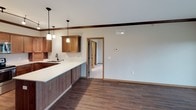 This single-story space is designed with functionality and cohesion in mind, incorporating warm wood tones and neutral finishes throughout. The kitchen is a standout feature, outfitted with rich medium-toned wood cabinetry, stainless steel appliances including a refrigerator, range, and over-the-range microwave, and light countertops. Track and pendant lighting illuminate the space, which also benefits from crown molding detail. A large island or peninsula with a white countertop and undermount sink extends into the open living area, which features dark hardwood-style flooring and exposed wood beam accents overhead. Pendant lights hang above the island, enhancing the open, connected feel between the kitchen and living spaces. The bedrooms are carpeted with neutral tones, each featuring windows that allow natural light to filter in. Two bathrooms serve the space: one includes a tub-shower combo with white surround, dark hardware accents, and a wood-cabinet vanity, while the second offers a walk-in shower and similar finishes. A dedicated laundry closet with stacked washer and dryer units adds convenience. Throughout, wood-trimmed door frames and warm beige walls create a consistent, welcoming aesthetic.