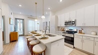 This well-appointed 2-bedroom, 2-bathroom residence showcases a bright, modern design with a cohesive neutral palette and quality finishes throughout its single floor. The open-concept kitchen serves as the heart of the home, featuring a large center island with a built-in sink and seating for multiple guests, illuminated by stylish gold pendant lights. White shaker cabinetry, a herringbone tile backsplash, stainless steel appliances—including a refrigerator, range, and microwave—and light wood-style flooring complete the space. The kitchen flows seamlessly into the living area, which benefits from natural light through sheer-draped windows and a glass door.

The primary bedroom is a generously sized retreat with plush carpeting, dual windows with sheer curtains, and access to a well-equipped en-suite bathroom featuring a dual-sink vanity with light wood cabinetry, a large mirror with globe lighting, and wood-look flooring. The second bedroom is equally comfortable with carpet flooring and natural light. Both bedrooms include dedicated closets, with the primary offering a walk-in closet with built-in shelving and hanging rods. The second bathroom features a tub-shower combination with classic white tile surround and a single-sink vanity.