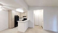 This single-floor space is designed with a practical, open layout that promotes easy movement between areas. The kitchen serves as a central feature, equipped with black appliances including a full-size refrigerator and a gas range with an overhead exhaust hood. Light wood cabinetry lines the walls, offering ample storage, while light-colored countertops provide functional workspace. A peninsula extends from the kitchen, creating a natural division between the cooking area and the adjacent living space, and offering additional counter space for casual use. The flooring in the kitchen and entryway area appears to be a neutral tile, while the adjoining space transitions to carpet, adding warmth underfoot. A textured ceiling is consistent throughout the space. The entryway, visible through an arched passage, is well-lit and leads to the exterior door, providing a welcoming transition point. Recessed and flush-mount lighting fixtures illuminate both the kitchen and the hallway, ensuring adequate brightness throughout. The overall color palette is neutral and understated, with white walls and light finishes creating a clean, airy atmosphere suited for everyday living.