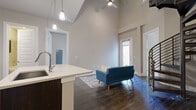 This single-floor residence is defined by its dramatic vaulted ceilings in the open-concept living and kitchen area, creating an airy, expansive atmosphere. Dark hardwood flooring flows throughout the main living spaces, complementing the neutral warm-gray wall tones. The kitchen is a standout feature, outfitted with rich dark-stained cabinetry—some with glass-front upper doors—light quartz countertops, a subway tile backsplash, and a full suite of stainless steel appliances including a refrigerator, range, and built-in microwave. A peninsula island with an undermount sink and pendant lighting above adds both function and style, seamlessly connecting the kitchen to the living area. The living space benefits from natural light filtering through a front entry door with sidelights, and a ceiling fan adds comfort overhead. Bedrooms feature plush carpeting; one includes a sliding glass door opening to an outdoor area. A generously sized walk-in closet with multiple shelving rods and rails provides ample storage. The bathroom is finished with large-format tile flooring, a tiled tub-shower surround, and dark vanity cabinetry consistent with the home's cohesive design aesthetic.