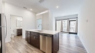This well-designed one-bedroom, one-bathroom unit is laid out on a single floor, offering an efficient and modern living environment. The open-concept kitchen and living area serves as the heart of the space, anchored by a sleek center island with a white quartz countertop, an undermount sink, and dark espresso cabinetry. The kitchen is equipped with a full suite of stainless steel appliances, including a refrigerator, range, built-in microwave, and dishwasher, complemented by a tile backsplash and recessed lighting overhead. Wood-look flooring runs throughout the entire unit, adding warmth and visual continuity. The living area benefits from abundant natural light, thanks to large sliding glass doors and floor-to-ceiling windows that offer open views of the surrounding streetscape and are dressed with neutral curtain panels. The bedroom is a calm, private retreat with clean white walls and ample room for furnishings, accessed through a paneled door. A dedicated closet provides convenient storage. The bathroom features a dark vanity with a white countertop, a framed mirror, and a glass-enclosed walk-in shower adorned with marble-style tile. The overall aesthetic is clean, contemporary, and cohesive throughout.