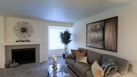 This single-story space presents a thoughtfully designed living area that balances comfort and contemporary style. The living room serves as the central gathering space, anchored by a fireplace that adds warmth and character to the room. A large window allows natural light to fill the space, brightening the neutral white walls and contributing to an open, airy atmosphere.

The flooring throughout the living area is fitted with plush carpet in a subtle, neutral tone, adding a layer of comfort underfoot. A glass-top coffee table sits at the center of the seating area, complementing a generously sized gray sofa accented with earth-toned throw pillows. The décor is cohesive and modern, highlighted by a large framed abstract artwork on the wall that serves as a visual focal point.

A potted indoor tree near the window introduces a natural element to the space, softening the clean lines of the interior. Additional decorative touches, including candle holders and a sculptural wall accent, round out the polished aesthetic. Overall, the space offers a calm, inviting environment with a neutral palette and carefully curated details throughout.