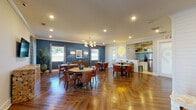 This contemporary communal area showcases a harmonious blend of style and functionality. The space is characterized by its striking blue accent walls, which provide a bold contrast to the white trim and ceiling. The flooring is a standout feature, with beautiful herringbone-patterned hardwood that adds warmth and sophistication.

The room is thoughtfully divided into multiple seating areas, each anchored by round wooden tables surrounded by comfortable, mid-century modern inspired chairs in rich brown tones. Overhead, modern globe chandeliers cast a warm glow, complementing the recessed lighting throughout the space.

Large windows allow natural light to flood the area, while stylish window treatments offer privacy when needed. The decor is carefully curated, with artwork adorning the walls and potted plants adding a touch of nature. A sleek kitchenette area is visible, featuring stainless steel appliances and ample counter space.

The space also includes a separate, glass-enclosed meeting room, perfect for more private gatherings or work sessions. This versatile area seamlessly combines elements of a lounge, dining room, and workspace, making it ideal for various social and professional activities. The overall design creates an inviting and dynamic environment that encourages interaction and community engagement.