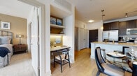 This single-floor residence showcases a thoughtfully designed open-concept layout that seamlessly connects the kitchen, dining, and living areas. The kitchen is outfitted with dark espresso cabinetry, stainless steel appliances, and a generous granite-topped island with pendant lighting overhead. Track lighting illuminates the dining area, where warm hardwood-style flooring flows throughout the main living spaces. A curated gallery wall of nautical-themed framed artwork adds character and visual interest to the dining space.

The living room is bright and welcoming, featuring large windows that invite natural light and offer outdoor views. A built-in nook near the entryway provides a dedicated workspace complete with granite countertop, under-cabinet lighting, and open shelving above — a practical and stylish addition to the layout.

The primary bathroom is elegantly finished with large-format tile flooring, a lengthy vanity featuring navy blue cabinetry, a wide mirror with Hollywood-style lighting, and direct access to the bedroom, which benefits from natural light through its own window. Throughout the residence, a neutral warm-toned palette with accents of navy and teal creates a cohesive and sophisticated atmosphere that balances comfort with modern design.