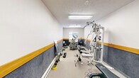 Located at 132 Andrew Pl in West Lafayette, Indiana, this dedicated fitness room offers a well-equipped workout space designed to support a range of exercise routines. The room features a distinctive two-tone wall treatment, with gray wainscoting along the lower half accented by a warm wood-tone trim band, topped by white upper walls — giving the space a clean, purposeful aesthetic. Bright fluorescent overhead lighting ensures the entire room is well-lit throughout. The floor is covered in a durable, textured surface suited for fitness activity.

The space is equipped with a variety of exercise machines and equipment, including a Reebok elliptical trainer, a recumbent stationary bike, and a multi-station cable weight system with an attached workout chart for reference. A power tower for bodyweight exercises stands against one wall, while a padded weight bench, dumbbells, weight plates, and a medicine ball provide additional free-weight and strength-training options. A full-length mirror leans against one wall, offering a practical aid for form-checking during workouts. A window along one wall allows in natural light and houses a wall-mounted AC unit. The room's compact but efficient layout makes it a fully functional facility for cardiovascular and strength training needs.
