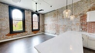 Located at 1277 W Sixth St in Cleveland's vibrant Ohio City neighborhood, this thoughtfully renovated space masterfully combines historic industrial character with contemporary design. Exposed brick walls — weathered to a rich patina of gray and amber — serve as a defining architectural feature throughout, creating a warm and textured backdrop against clean white finishes and dark hardwood floors.

The open main area features a modern kitchen equipped with stainless steel appliances, white cabinetry, a sleek countertop island, and elegant pendant lighting. Large arched and floor-to-ceiling black-framed windows flood the space with natural light while offering views of the surrounding streetscape. A ceiling fan adds a functional, loft-inspired touch overhead.

A private room provides a quieter retreat, featuring soaring ceilings with a clerestory window that draws in additional light, complemented by the same rich hardwood flooring found throughout. A second room showcases more of the signature exposed brick alongside modern white-paneled doors and black hardware.

The hallway is clean and well-lit, with exposed ductwork adding an industrial accent above. The bathroom is a standout, pairing the exposed brick accent wall with a white vanity, matte black fixtures, a large mirror, and a glass-enclosed walk-in shower dressed in marble-style tile. Every detail reflects a deliberate balance of historic character and refined, modern living.