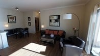 This single-floor space showcases an open-concept layout that connects the living and dining areas, creating a cohesive and functional environment. The rich dark hardwood flooring runs throughout, providing a warm and grounding aesthetic that complements the neutral wall tones.

The living area is anchored by a dark leather sofa accented with terracotta and rust-toned throw pillows, adding a pop of color against the understated backdrop. A sleek arc floor lamp provides ambient lighting, while a rectangular coffee table with a chrome frame serves as a central focal point. A bold abstract framed artwork adorns the main wall, contributing a contemporary artistic touch.

Natural light streams in from windows near the dining area, where a dark dining table and chairs offer a dedicated space for meals. Crown molding trims the ceiling throughout, adding a refined architectural detail that elevates the overall finish of the space.

A hallway visible from the main living area leads to additional rooms, suggesting a thoughtfully designed floor plan. The overall ambiance is modern, warm, and inviting, with a consistent color palette that ties each area together seamlessly.