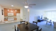 This single-floor residence features an open-concept layout that seamlessly connects the kitchen, dining, and living areas, creating a cohesive and functional living environment. The kitchen is equipped with warm honey-toned wood cabinetry, a white refrigerator, a stove, and a double sink set into a light-colored countertop. A peninsula counter with pendant lighting above serves as a natural divider between the kitchen and the dining area, offering additional prep or casual seating space. The dining area accommodates a dark wood table with matching cross-back chairs and is centered beneath a ceiling fan with an integrated light fixture, which also helps circulate air throughout the open space. The adjoining living area features neutral-toned carpeting that adds warmth underfoot and provides ample room for a media setup. The walls throughout are painted in a clean, light neutral tone, contributing to a bright and airy atmosphere. The open flow between all three areas makes the space feel expansive and well-connected, ideal for both everyday living and casual entertaining. The single-floor layout ensures easy accessibility and a straightforward, practical floor plan.