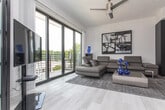 This contemporary single-floor residence showcases a thoughtfully designed open-concept layout with clean lines and a modern aesthetic throughout. The living area is defined by wood-look flooring, a ceiling fan, and floor-to-ceiling sliding glass doors that open to a private balcony with outdoor views. A striking frosted glass and black-framed sliding partition elegantly separates the living space from the bedroom, allowing natural light to pass through while providing privacy when needed.

The bedroom accommodates a full-size bed and includes a ceiling fan, a window with exterior views, and direct visual connection to the balcony through the glass partition. The galley-style kitchen is well-appointed with high-gloss white cabinetry extending to the ceiling, stainless steel appliances including a gas range, built-in microwave, dishwasher, and refrigerator, complemented by quartz countertops and a neutral tile backsplash.

The bathroom features large-format grey tile flooring, a floating vanity with an undermount sink and grey countertop, a wall-mounted toilet, and a glass-enclosed walk-in shower with a rain showerhead. The overall ambiance is clean, minimal, and contemporary.
