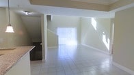 This two-story space showcases an open-concept layout that maximizes both functionality and natural light. The main living area features large-format tile flooring that extends throughout, providing a clean, cohesive look underfoot. A generous sliding glass door fitted with vertical blinds serves as a focal point of the living space, flooding the interior with abundant natural light and offering access to the outdoors.

The soaring ceiling height creates a voluminous, airy atmosphere, enhancing the sense of spaciousness throughout the main level. A staircase positioned within the open-plan layout provides access to the upper floor, while a structural column adds architectural definition to the space without interrupting its open flow.

To one side, a partial view of the kitchen reveals dark cabinetry and a granite or stone countertop, suggesting a well-appointed culinary space adjacent to the main living area. The walls are finished in a neutral, light tone that complements the overall brightness of the space. White baseboards provide clean contrast against the flooring, contributing to the polished, well-maintained character of this two-level home.