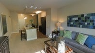 This single-story residence showcases an open-concept layout that connects the living, dining, and kitchen areas into one cohesive, functional space. The living area is carpeted and comfortably sized, accommodating a plush sofa, glass-top coffee table, and accent furnishings, with natural light streaming in to create a bright and welcoming atmosphere. A contemporary piece of wall art adds a pop of color to the neutral-toned walls.

The kitchen is well-appointed, featuring dark cabinetry, black appliances including a full-size refrigerator, and a granite-topped island with seating — ideal for casual dining or entertaining. Pendant lighting hangs above the island, adding both style and task lighting to the space. The kitchen flows naturally into a dedicated dining area, which is illuminated by an overhead light fixture and accented with greenery.

Hardwood-style flooring runs through the kitchen and dining zones, transitioning to carpet in the living area, giving each space a distinct feel while maintaining visual continuity throughout. The overall design balances practicality with modern aesthetics.