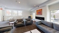 The clubhouse at 703 Whitney Ave in New Haven, Connecticut, is a thoughtfully designed single-floor community gathering space that blends modern aesthetics with comfortable, functional living. The open-concept layout is anchored by a generous lounge area furnished with multiple gray sofas and oversized armchairs, all arranged around a marble-top coffee table and an accent side table in warm wood tones. A sleek, linear fireplace runs along one wall, adding warmth and a striking architectural focal point to the room, while a large wall-mounted flat-screen TV and a contemporary media console with wood-drawer detailing provide entertainment options. Vibrant abstract artwork in bold oranges, reds, and blues punctuates the crisp white walls, lending the space a curated, gallery-like quality. Patterned carpeting in deep blue-gray tones adds texture underfoot and defines the seating zones throughout. A separate dining area features round white tables paired with Eames-style chairs, bathed in natural light streaming through a generous bank of windows that frame views of the surrounding neighborhood. Recessed lighting throughout ensures a bright, welcoming atmosphere at any time of day. The overall ambiance is clean, contemporary, and inviting — ideal for residents seeking a well-appointed shared space for socializing or relaxing.