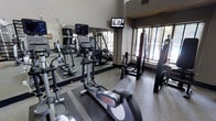 This fitness center is designed to support a comprehensive workout experience within a compact, efficiently arranged space. The room features soaring ceilings that lend an open, airy feel, preventing the space from feeling cramped despite its focused footprint. Large windows along one wall invite natural light and provide views of outdoor greenery, creating an energizing atmosphere for exercise.

The cardio section is anchored by Life Fitness elliptical trainers and treadmills, positioned for easy use and visibility. A wall-mounted flat-screen television is positioned at an optimal viewing angle, allowing users to stay entertained during workouts. Full-length mirrors line one wall, enhancing the sense of depth and allowing users to monitor their form during exercise.

Strength training is supported by a multi-functional cable and weight machine, rounding out the equipment offerings for a balanced fitness routine. The flooring consists of durable, rubber-textured material suited for high-traffic athletic use. The overall ambiance is clean and functional, with neutral tones and practical lighting contributing to a focused, motivating environment ideal for daily fitness routines.