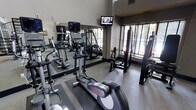 This fitness center is designed to support a comprehensive workout experience within a compact, efficiently arranged space. The room features soaring ceilings that lend an open, airy feel, preventing the space from feeling cramped despite its focused footprint. Large windows along one wall invite natural light and provide views of outdoor greenery, creating an energizing atmosphere for exercise.

The cardio section is anchored by Life Fitness elliptical trainers and treadmills, positioned for easy use and visibility. A wall-mounted flat-screen television is positioned at an optimal viewing angle, allowing users to stay entertained during workouts. Full-length mirrors line one wall, enhancing the sense of depth and allowing users to monitor their form during exercise.

Strength training is supported by a multi-functional cable and weight machine, rounding out the equipment offerings for a balanced fitness routine. The flooring consists of durable, rubber-textured material suited for high-traffic athletic use. The overall ambiance is clean and functional, with neutral tones and practical lighting contributing to a focused, motivating environment ideal for daily fitness routines.