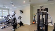 Located at 31611 Plaza De Toros Dr. in Spanish Fort, Alabama, this single-floor fitness space is thoughtfully designed to support a well-rounded exercise routine. The room features neutral, light-toned walls accented with crisp crown molding and white baseboards, giving the space a clean and polished aesthetic. Recessed lighting in the ceiling provides consistent, even illumination throughout, while a ceiling fan adds comfort during workouts. The flooring is covered in a low-pile carpet suited for the fitness environment. Multiple wall-mounted flat-screen televisions are positioned for easy viewing from various workout stations, enhancing the exercise experience. The space is equipped with a range of fitness machines, including an elliptical trainer and a stationary upright bike, as well as a multi-function cable and weight machine for strength training. A collection of medicine balls and other accessories are neatly arranged in the corner, offering options for varied training routines. The overall ambiance is practical and well-maintained, reflecting a dedicated approach to wellness with a layout that maximizes usability while maintaining an orderly and inviting environment.