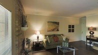 This single-floor living space centers around a well-appointed living room that balances comfort and style. A prominent stone fireplace anchors one side of the room, adding a classic architectural element and a natural focal point. The fireplace mantel provides display space, currently accented with decorative greenery and basket accents.

The living room features warm-toned walls complemented by crown molding that adds a refined finishing detail. Dark hardwood flooring grounds the space and contrasts attractively with the light walls. The ceiling carries a gentle slope, contributing to the room's sense of character.

Furnishings include a plush, earth-toned sofa adorned with decorative throw pillows in complementary colors, paired with a sleek glass-top coffee table that keeps the space feeling open. A matching glass-top end table holds a classic table lamp, casting warm ambient light throughout the room. Wall art, including a landscape-inspired canvas and a set of decorative wall panels, adds visual interest without overwhelming the space.

Overall, the living area presents a warm, inviting ambiance suited for relaxed everyday living, with thoughtful details throughout.