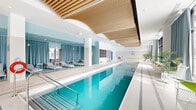 Located at 1240–1250 Cummings Ave in Ottawa, Ontario, this elegantly designed indoor pool facility spans a generous single floor, offering a resort-inspired aquatic environment. At its centre sits a pristine lap pool with crystal-clear turquoise water, framed by fine white mosaic tile decking that extends throughout the space. Stainless steel entry rails provide easy pool access, while safety equipment is thoughtfully positioned along the perimeter.

The ceiling is a standout architectural feature — a sculpted installation of warm wood slats that undulates gracefully above the pool, adding natural texture and visual depth against the bright white surroundings. Floor-to-ceiling windows line one side of the space, dressed with flowing teal curtains that can be drawn for privacy or opened to flood the room with natural light and outdoor views of the Ottawa cityscape.

A dedicated lounge area furnished with contemporary chaise lounges, wicker chairs, and side tables provides a comfortable retreat for relaxation before or after swimming. Potted tropical palms and greenery are distributed throughout, enhancing the spa-like atmosphere. Built-in storage shelving and safety equipment are neatly integrated along the walls. The overall design balances clean, modern minimalism with warm, welcoming details, creating a sophisticated and functional aquatic amenity.