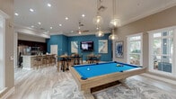 This single-story amenity space is thoughtfully designed to accommodate social gatherings and recreation. The grand entrance features a striking arched transom window flanked by tall panel windows and French doors, flooding the foyer with natural light and creating an open, welcoming atmosphere. Light-toned hardwood-style flooring and crown molding run throughout, lending a cohesive, polished aesthetic.

The main gathering area is anchored by a bold teal accent wall and includes a wall-mounted flat-screen television, making it ideal for communal viewing. Multiple high-top tables with swivel bar stools and coastal-themed artwork create a relaxed yet refined dining lounge atmosphere. A separate seating area features upholstered armchairs arranged around a central coffee table beneath a cluster of globe pendant lights.

The game room zone showcases a full-size billiards table positioned beneath additional pendant lighting, complemented by a foosball table visible in an adjoining area.

The kitchen is equipped with stainless steel appliances, dark cabinetry, and a large marble-topped bar island with seating, ideal for casual entertaining. Ceiling fans, recessed lighting, and an abundance of windows ensure the space remains bright and comfortable throughout.